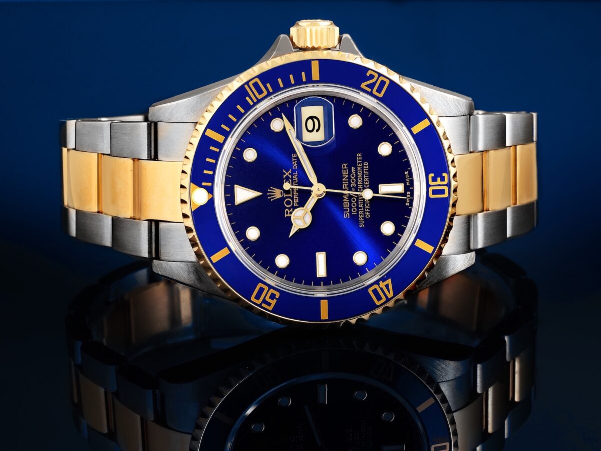 A sharp close-up studio photograph of a two-tone Rolex Submariner Reference 16613 ("Bluesy") with a cobalt blue sunburst dial and blue bezel, on a deeply reflective dark surface.