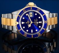 A sharp close-up studio photograph of a two-tone Rolex Submariner Reference 16613 ("Bluesy") with a cobalt blue sunburst dial and blue bezel, on a deeply reflective dark surface.