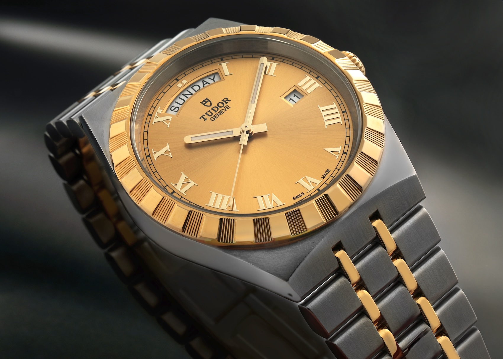 igh angle view of a Tudor Royal watch with two-tone steel and yellow gold notched bezel, integrated bracelet, champagne dial, and black Roman numerals.