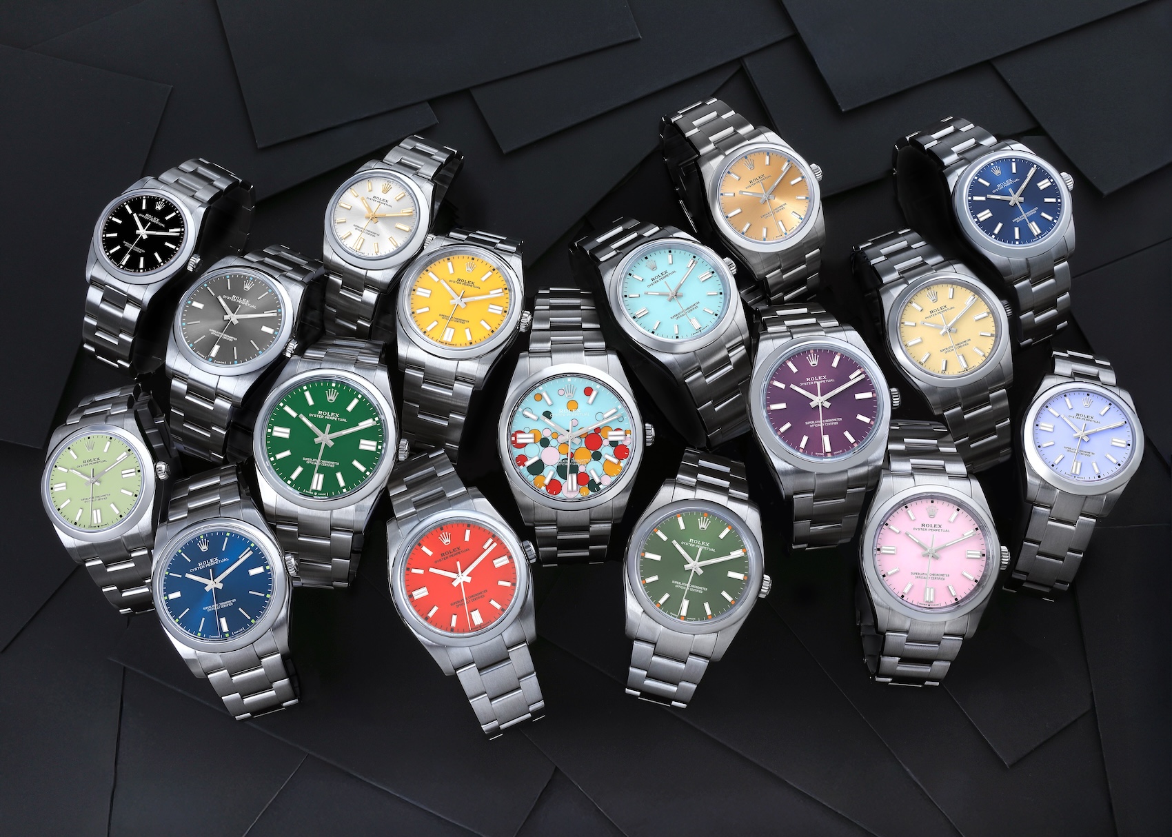 An overhead flat lay photograph of fifteen Rolex Oyster Perpetual watches featuring varied colorful dials arranged on a textured black background, with the specific "Celebration" bubble dial prominent in the center.