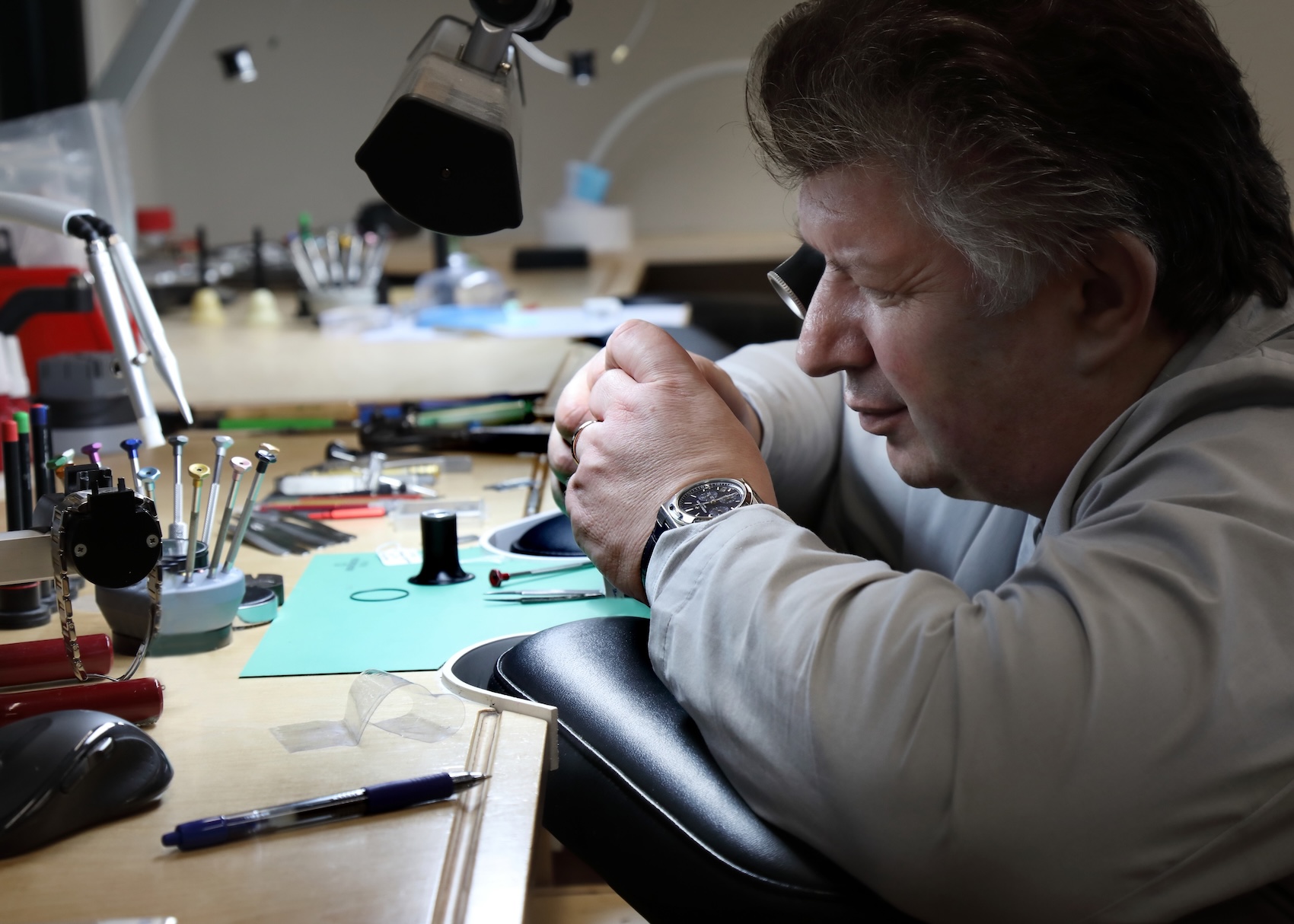 An expert watchmaker at SwissWatchExpo using a loupe at a workbench to carefully inspect an open mechanical watch movement.