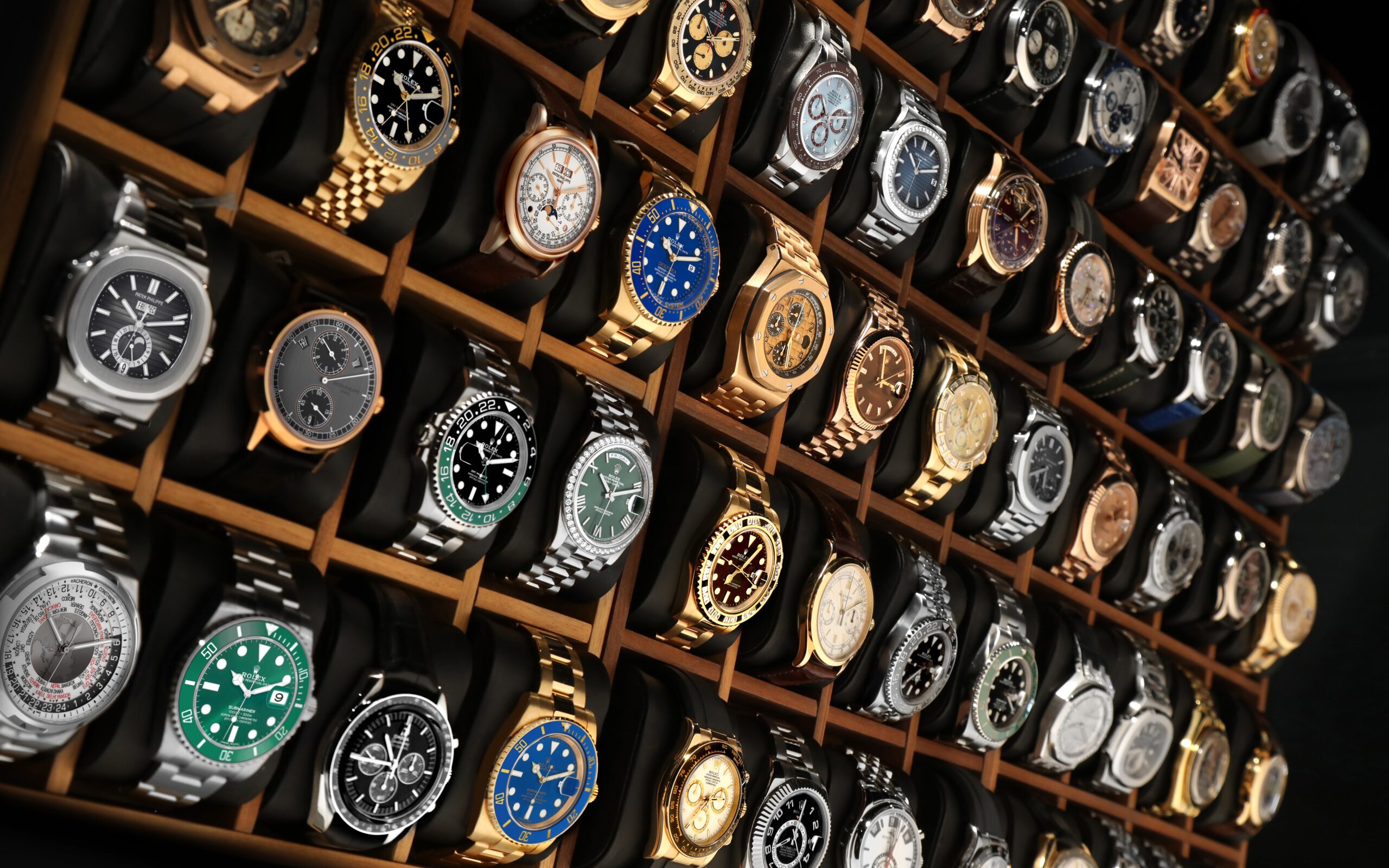 A large wooden display case filled with rows of pre-owned luxury watches, including models from Rolex, Patek Philippe, and Audemars Piguet.
